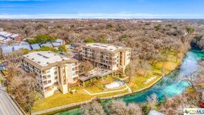 A multi-story building with a flat roof, surrounded by a river and trees. - Updated Single Story Comal River Condo with Pool-Walk to Downtown NB (New Braunfels)
