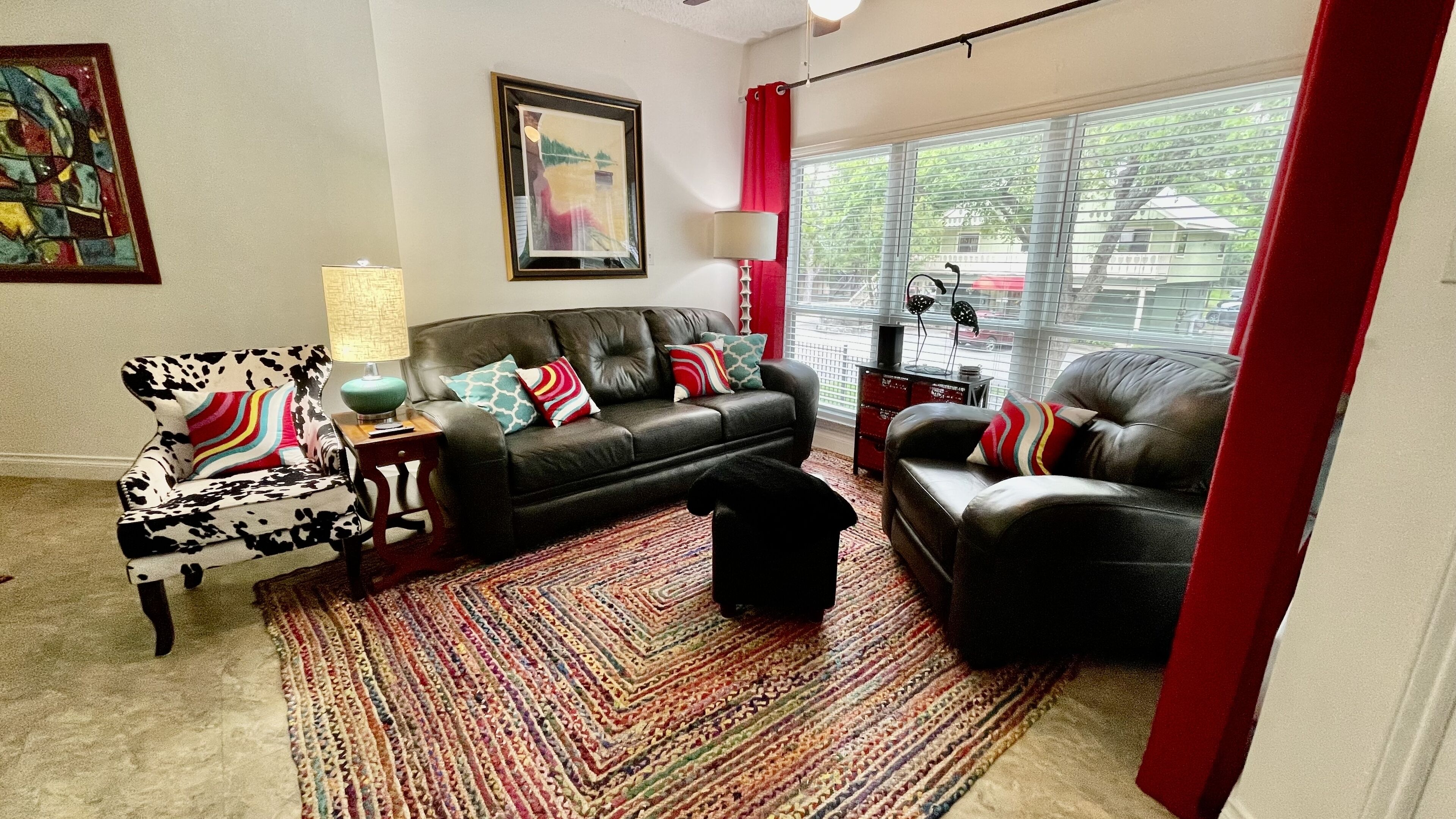A living room with a black leather sofa, a patterned armchair, a colorful rug, and a large window with blinds.