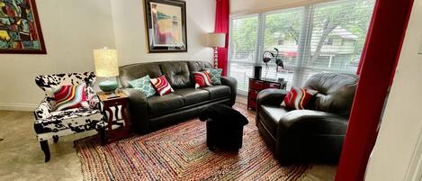 A living room with a black leather sofa, a patterned armchair, a colorful rug, and a large window with blinds.