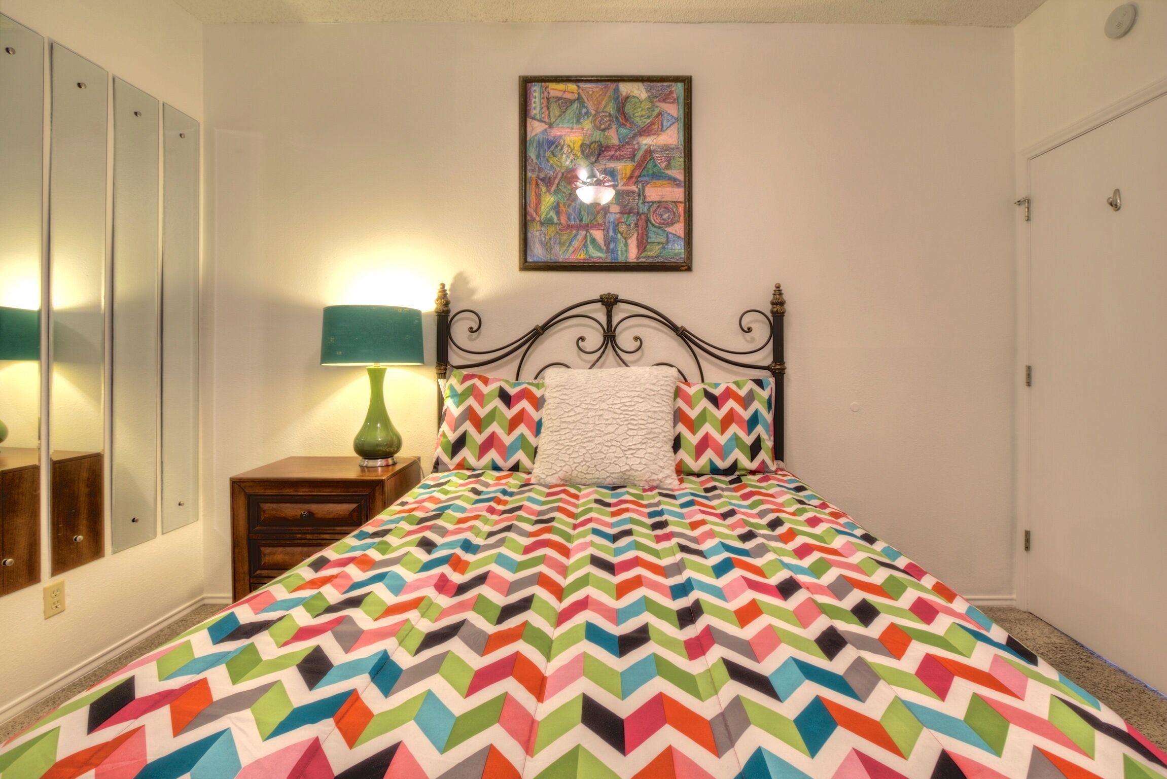Un dormitorio con una cama, una mesita de noche, una lámpara, un espejo y un cuadro de colores en la pared.