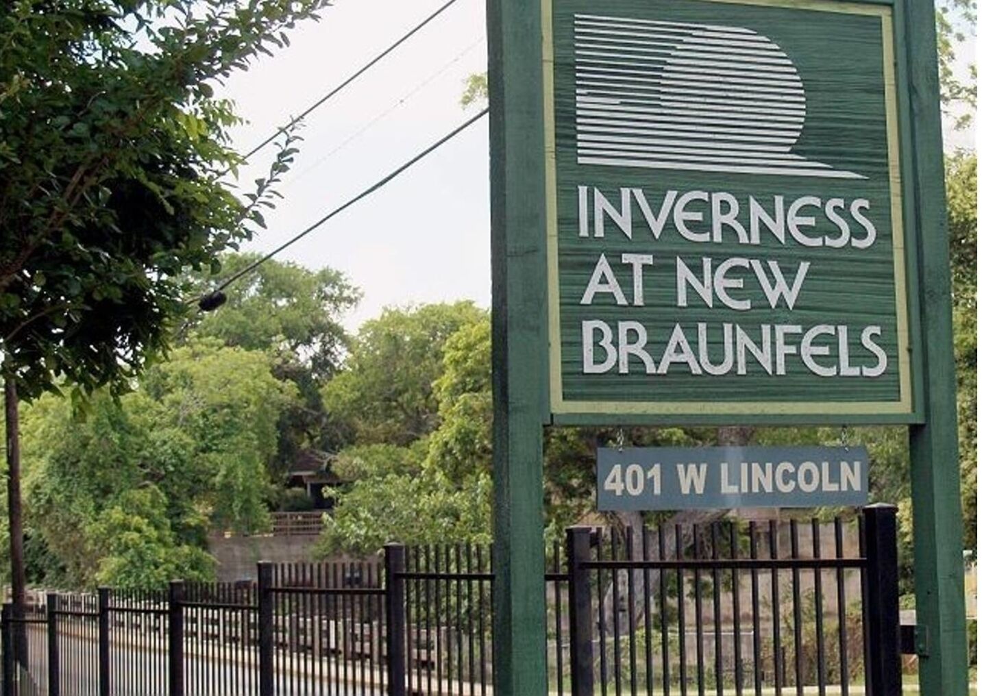 En skylt med texten ”Inverness at New Braunfels”, där man delvis ser ett staket och träd i bakgrunden.