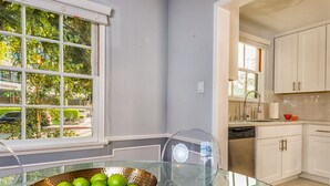 A modern kitchen with white cabinets, a glass dining table with a copper bowl of fruit, and a view of the outside through a large window. - City Escape Living (Burbank)