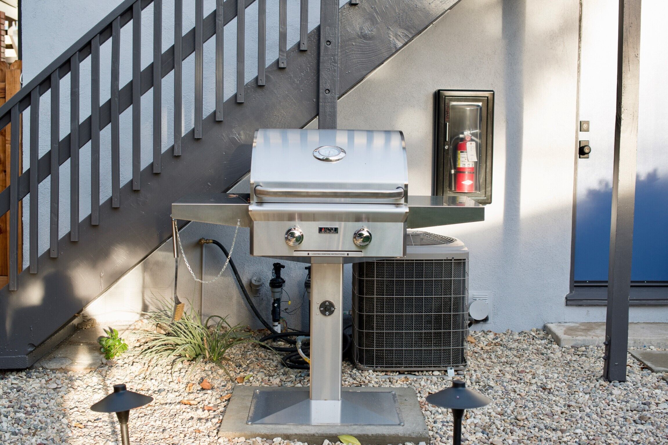 An outdoor grill setup with a fire extinguisher, an air conditioning unit, and a staircase.