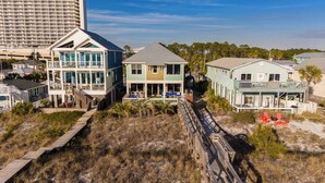 A beachfront property with a wooden boardwalk leading to a multi-story building with a balcony, a separate building with a porch, and a light blue house with a deck.