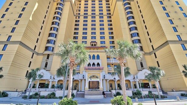 Un hotel gran de diversos pisos amb una façana simètrica, que presenta una entrada central amb un arc decoratiu i palmeres al davant.