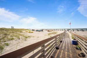 Ein hölzerner Steg führt zu einem Strand mit Sonnenschirmen und Liegestühlen.