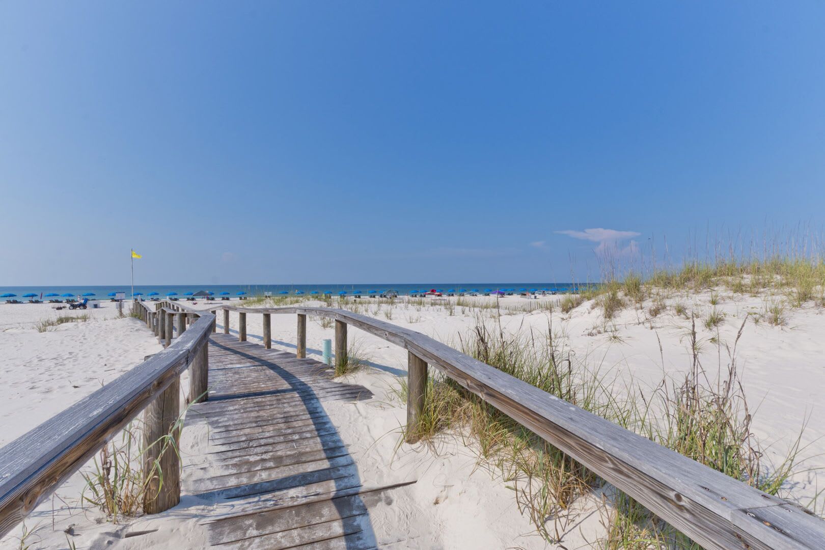 A wooden boardwalk leading to a beach with sand dunes and a clear blue sky.