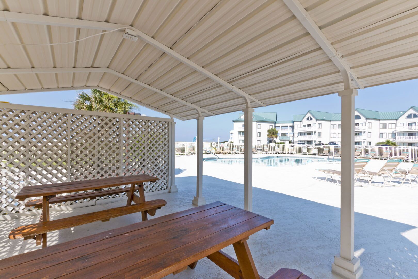 A covered outdoor area with a wooden picnic table, a pool, and a view of hotel buildings in the background.