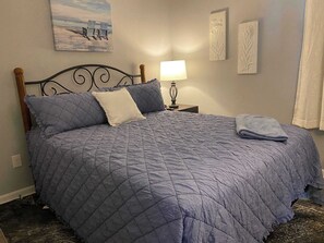 Ein ordentlich bezogenes Bett mit einer blauen Steudecke, einer Nachttischlampe und einem Bild von Strandstühlen an der Wand.