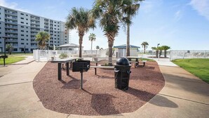 A park area with a barbecue grill, picnic tables, and a trash can.