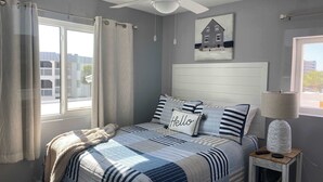 A bedroom with a bed, a ceiling fan, a bedside lamp, a window with curtains, and a painting of a house.