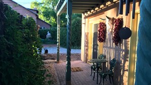 Un espacio al aire libre techado con una mesa y sillas, una puerta adornada con coronas decorativas y una vista de una casa al fondo.
