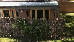 Una estructura de madera rústica con techo inclinado, posiblemente un cobertizo o zona de almacenamiento, rodeada por una valla de madera y vegetación.