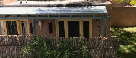 Una estructura de madera rústica con techo inclinado, posiblemente un cobertizo o zona de almacenamiento, rodeada por una valla de madera y vegetación.