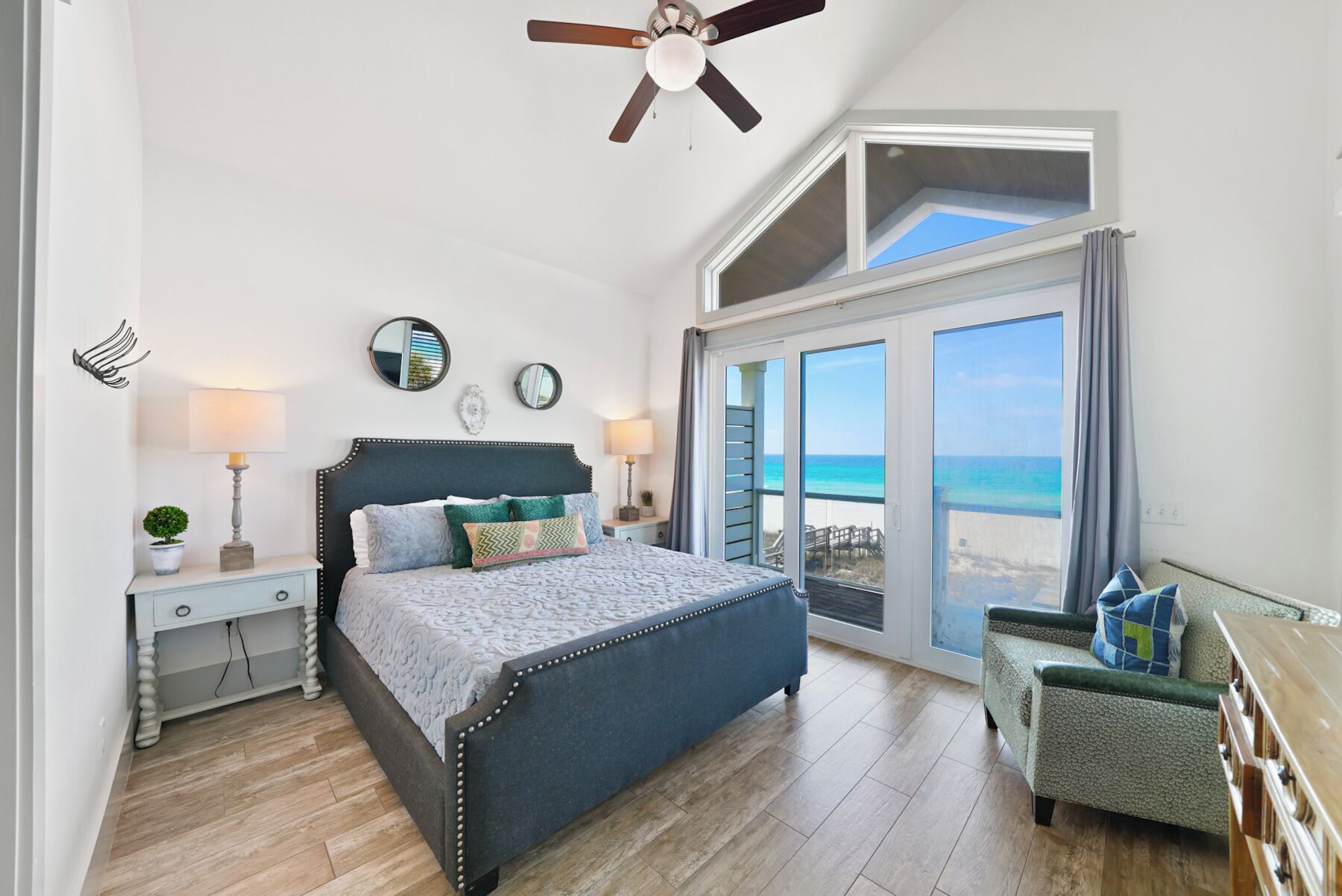 Ein Schlafzimmer mit einem großen Bett, einem Deckenventilator, einem Nachttisch mit Lampe, einem Schminktisch und Blick auf den Strand durch eine Glasschiebetür.