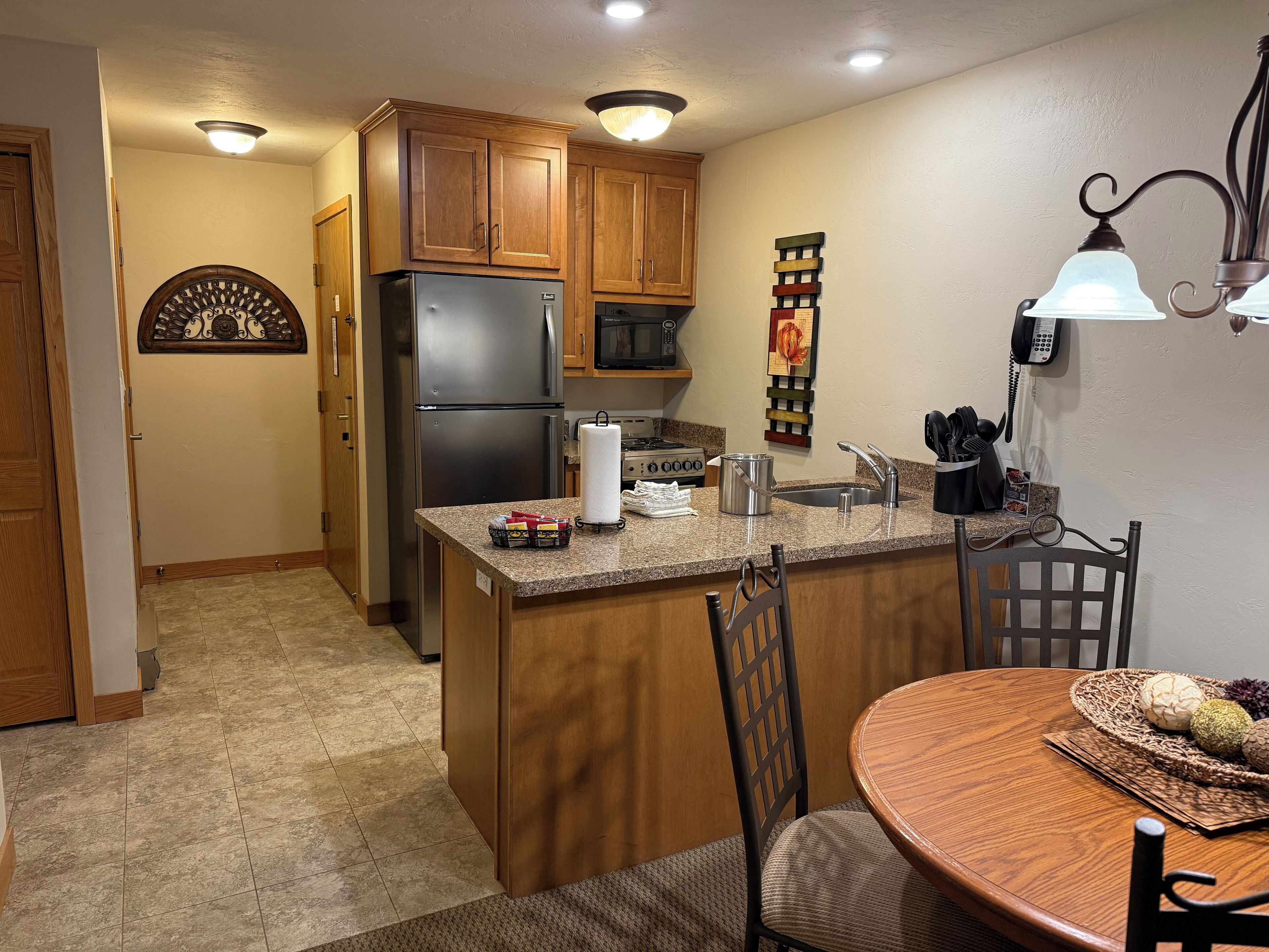 A kitchen with wooden cabinets, a stainless steel refrigerator, a microwave, and a stove. There is a dining table with chairs and a decorative wall hanging.