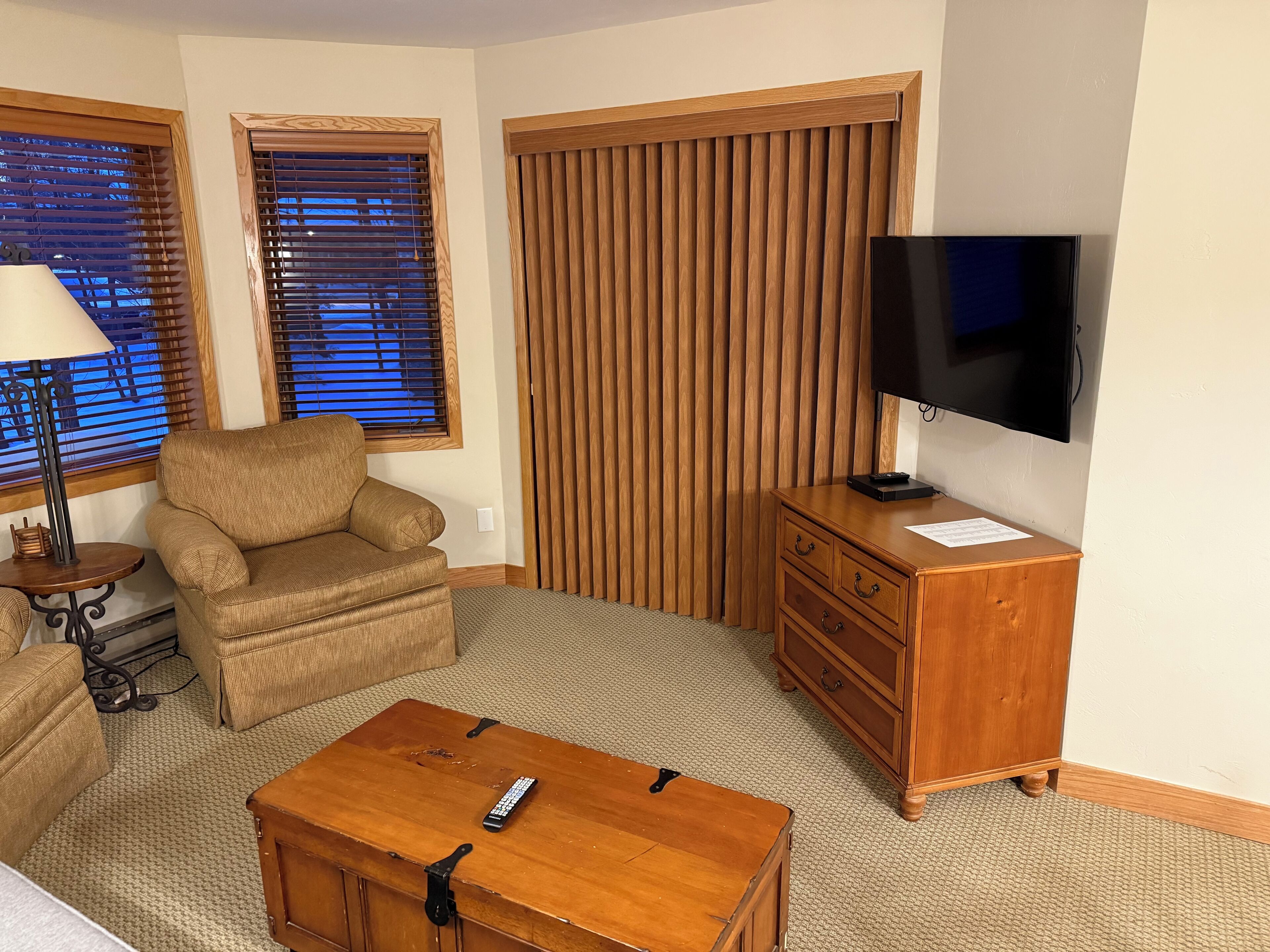 Una sala de estar con sofá, sillón, mesa de centro de madera, mueble para televisor y persianas.