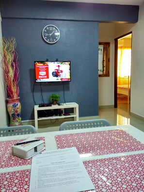 A hotel room with a TV on the wall, a clock, a desk with a computer, and a bed with a patterned bedspread. - iCheck Homestay Putrajaya (Putrajaya)
