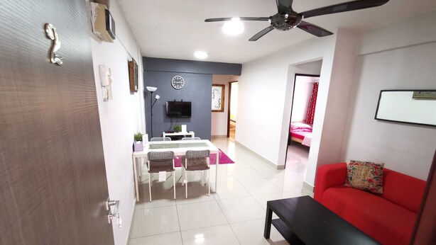 A living room with a red sofa, a black coffee table, a dining area with a white table and chairs, a ceiling fan, and a TV mounted on the wall. - iCheck Homestay Putrajaya (Putrajaya)