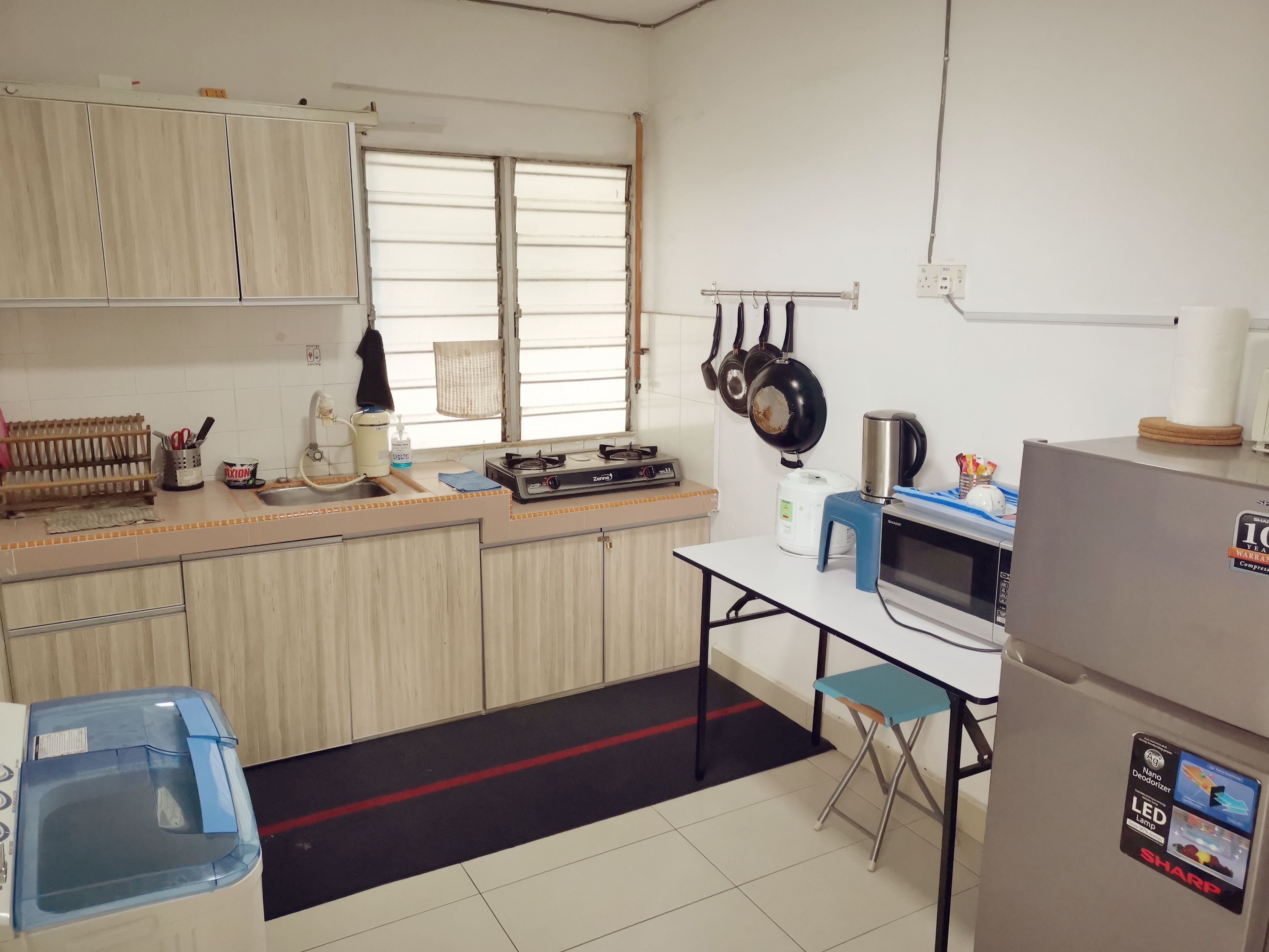 A compact kitchen with a gas stove, microwave, and a refrigerator. There is a sink with a faucet, a window with blinds, and a small stool.