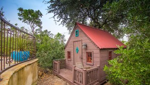A small wooden cabin with a red roof, a porch with a chair, and a metal fence with a blue object.