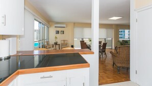 Una cocina moderna con encimera negra y armarios blancos, que da paso a un comedor con sillas de mimbre y vistas a la ciudad.