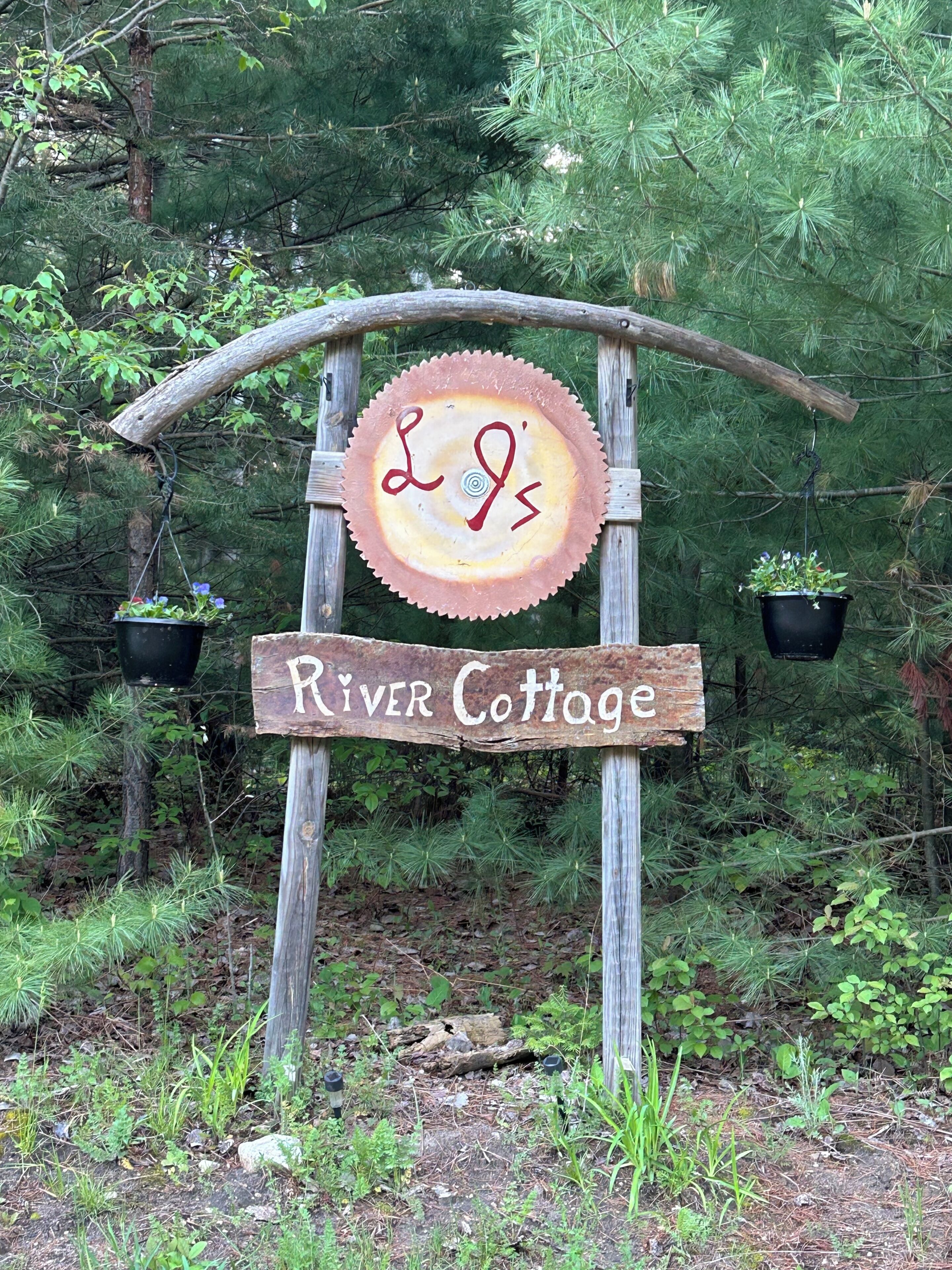 Un cartello di legno rustico con su scritto 'River Cottage', sostenuto da un arco di legno, con vasi pensili da entrambi i lati.