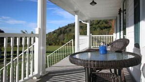 A covered porch with a round table and wicker chairs, white railings, and a view of a green landscape. - Awesome Flat Top Farmhouse - Ashwood Rentals (Flat Top)