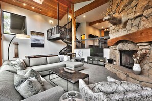 A modern living room with a stone fireplace, a large sectional sofa, a wooden staircase, and a flat-screen TV. - Big 5 Bed Home Lower DV, Hot Tub, Pool Table, AC, DV Ski Shuttle! Up to 25% Off! (Park City)