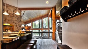 Un interior rústico con pared de piedra, vigas de madera, un ventanal grande y una zona de bar con taburetes.