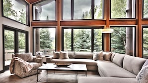 Una sala de estar moderna con un sofá seccional, una chaise longue y una mesa de centro. Ventanas amplias ofrecen vistas a un área boscosa.