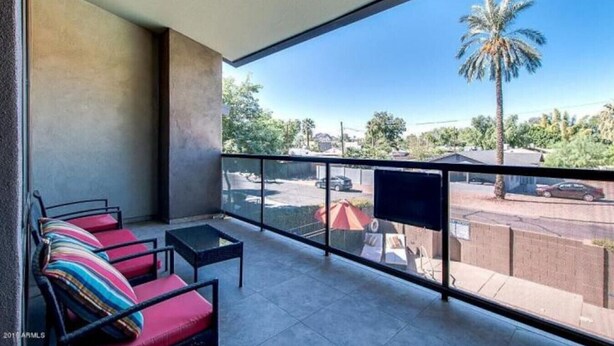A balcony with a sofa and chairs, a TV screen, and a view of a street with palm trees and parked cars.