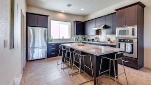 A modern kitchen with dark cabinetry, stainless steel appliances, a large island with bar stools, and a window providing natural light.
