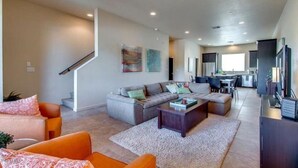 A spacious living room with a grey sectional sofa, orange armchairs, and a coffee table. A staircase leads to an upper level.
