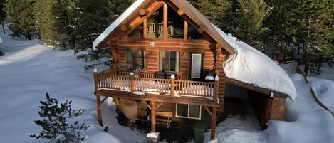 Une cabane en rondins dotée d’un balcon et d’un toit enneigé, entourée d’arbres.