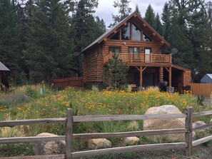 Une cabane en rondins dotée d’un balcon, entourée d’une clôture en bois et d’un champ de fleurs jaunes.