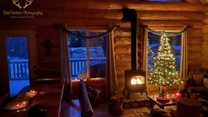 Interior acogedor de una cabaña con pared de troncos, chimenea, un árbol de Navidad y vistas a una terraza nevada.