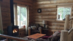Interior acogedor de una cabaña de madera con estufa de leña, un sofá, una mesa y una ventana con vistas a una terraza.