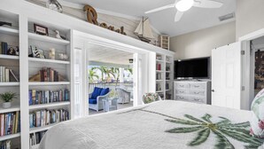 A bedroom with a large bed, a TV, and a bookshelf. There is a balcony with a view of palm trees. - Pool, Spa, Kayaks, Bikes, Fishing Gear & More waterfront, Amenities at no charge (Cape Coral)