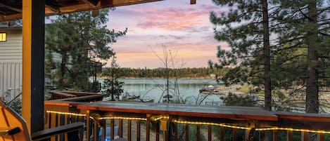 A wooden deck with chairs and string lights, overlooking a lake with trees and a dock.