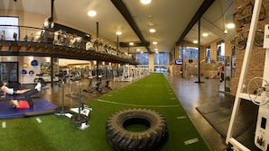 Une salle de sport équipée de divers appareils d’exercice, avec une piste verte qui la traverse en son centre et un mur de briques sur lequel sont accrochés des vélos, sur le côté droit.