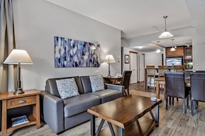 A modern living room with a sofa, coffee table, and lamps. There is a dining area with chairs and a table. A kitchen is visible in the background. - Relax & Explore: Mountain Views, Heated Pool & Dog-Friendly Condo (Canmore)