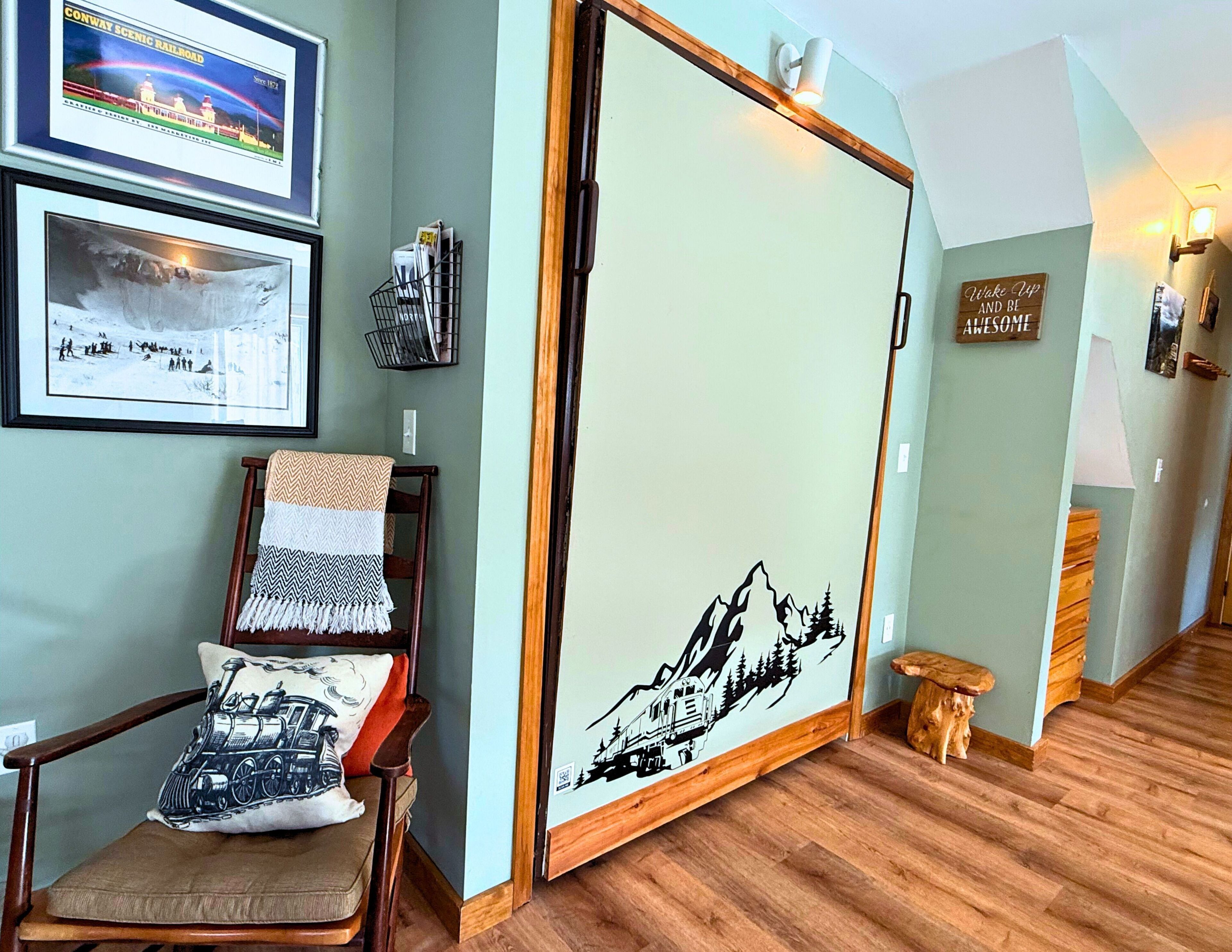 Une pièce avec un plancher en bois, une chaise dotée d’un coussin à motifs, un mur orné d’un décor représentant un paysage de montagne et un tableau encadré au-dessus de la chaise.