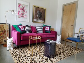 Un salon avec un canapé rouge, deux chats, un tapis à motifs et divers objets décoratifs, notamment une lampe et des œuvres d’art encadrées.