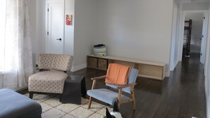 Ein Wohnzimmer mit einer Couch, einem Sessel, einer Bank und einem kleinen Tisch. Eine Tür und ein Flur führen in einen anderen Raum.
