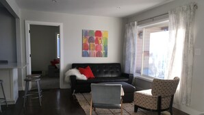 Ein Wohnzimmer mit einem schwarzen Ledersofa, einem gemusterten Sessel, einem Couchtisch und einem Teppich. An der Wand hängt ein Gemälde und ein Fenster ist mit Vorhängen drapiert.
