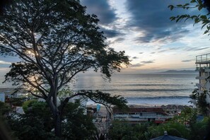 Une vue côtière avec un arbre au premier plan, une plage et un bâtiment doté d’un balcon donnant sur la mer.