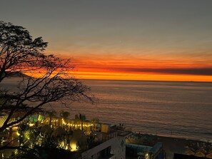 A coastal view with a sunset, a tree in the foreground, and a lit-up resort area with umbrellas.