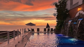 Área da piscina com vista para o pôr do sol, cadeiras de descanso e uma cascata.