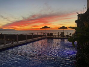 A poolside view with lounge chairs and umbrellas, overlooking a sunset over the ocean.